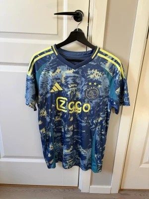 Ajax blå matchtröja Adidas - Säljer en blå Ajax matchtröja från Adidas med coolt mönster i blått och gult. Tröjan har gula detaljer på axlarna och logotypen Ziggo på bröstet. Materialet är lätt och andas, perfekt för fotboll eller träning. Ajax-emblem med tre stjärnor på framsidan.
