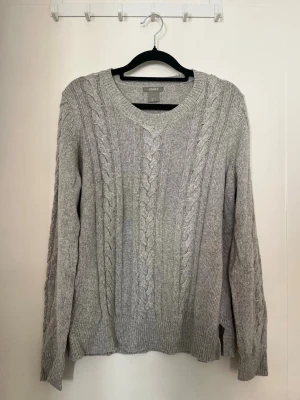 Grå stickad tröja - Snygg grå kabelstickad tröja från Lindex med v-ringning och långa ärmar. Tröjan har klassiska flätmönster framtill och på ärmarna samt ribbade muddar vid ärmslut och nederkant. 