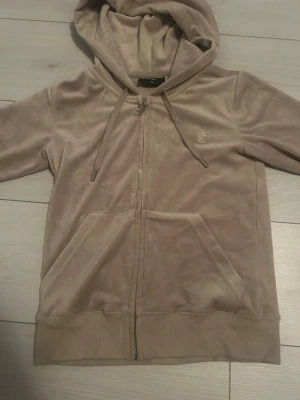 Juicy Couture beige hoodie - Säljer en beige hoodie från Juicy Couture med dragkedja, huva och snören. Tröjan har två stora fickor framtill och ett litet JC-logo broderat på bröstet. Mjukt material och ribbade muddar vid ärmslut och nederkant. 💕
