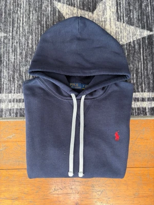 Mörkblå Polo Ralph Lauren Fleece Hoodie - Storlek M - Hej! 👋 Säljer en riktigt snygg mörkblå (Navy) Polo Ralph Lauren hoodie i storlek M. Den har den klassiska röda pony-loggan broderad på bröstet 🐎 Tröjan är i mycket gott skick och välvårdad ✨ Den är 100% äkta med både tvättlappar och QR-kod kvar för enkel verifiering ✅ Ett tidlöst premium-basplagg som passar till allt och håller hög kvalitet år efter år. Rekommenderas starkt! 👕 Fraktar snabbt! 🚛📦💬💯 