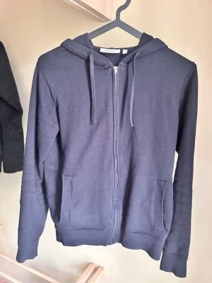Mörkblå zip-hoodie med huva - Jag är 165. En mörkblå hoodie från New Yorker med dragkedja, huva och snören. Tröjan har två fickor framtill och ribbade muddar vid ärmslut och nederkant. Perfekt för en avslappnad och cool look. Materialet är mjukt och bekvämt, troligtvis bomullsblandning.