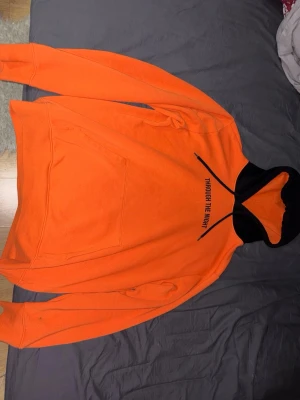 Orange hoodie med svart huva, Storlek M/L - Cool orange hoodie med svart huva och texten 'Through The Night' på bröstet. Hoodien har en stor magficka och svarta snören vid huvan. Perfekt för dig som vill sticka ut och gillar streetwear-stilen.