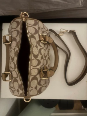 Handväska med logomönster - Snygg beige handväska  med det klassiska mönstret i brunt. Väskan har både handtag och avtagbar axelrem i läder, samt guldfärgade metalldetaljer. Perfekt storlek för det viktigaste och stängs med dragkedja.