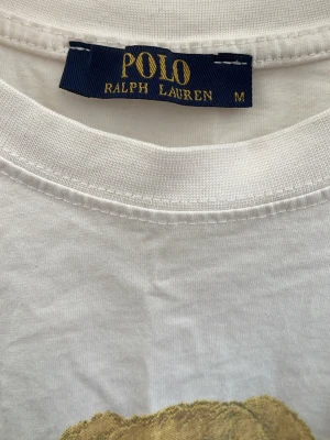 Vit Polo Bear t-shirt från Ralph Lauren - Vit t-shirt från Ralph Lauren med ikoniskt Polo Bear-tryck framtill. Klassisk rund hals och korta ärmar. T-shirten är i mjuk bomull och har en avslappnad passform. Perfekt för dig som gillar streetstyle och vill ha en tidlös look. Inget kvitto.