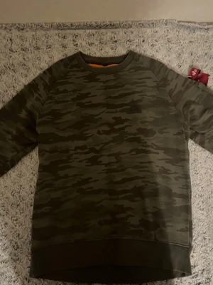 Bra dröja prist kan diskuteras inga fel med den - Snygg sweatshirt i militärgrönt camomönster från Alpha Industries. Tröjan har rund halsringning, ribbade muddar och en klassisk passform. Perfekt för dig som gillar streetwear och vill ha en avslappnad look.