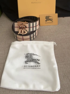 Burberry rutigt bälte med guldspänne - Säljer ett klassiskt Burberry bälte med ikoniskt rutigt mönster i beige, svart, rött och vitt. Bältet har ett stort guldigt B-spänne framtill och är tillverkat i skinn. Perfekt accessoar för att lyfta vilken outfit som helst.