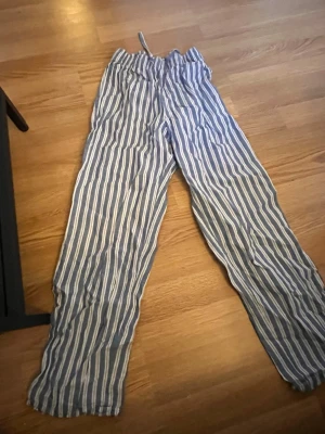Randiga pyjamasbyxor i bomull - Säljer ett par blåvita pyjamasbyxor med vertikala ränder och dragsko i midjan. Byxorna är löst sittande och tillverkade i ett mjukt bomullstyg, perfekta för att chilla hemma eller sova i.