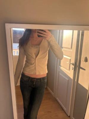 Beige ribbad kofta från Gina Tricot - Säljer en beige ribbad kofta från Gina Tricot i storlek XS. Koftan har långa ärmar, v-ringning och knäppning framtill med små vita knappar. Perfekt att styla med ett linne under för en enkel och snygg look.