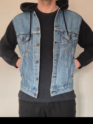 Levi's jeansjacka hybrid med huva - Snygg jeansjacka från Levi's med svarta ärmar och huva i sweatshirtmaterial. Klassisk blå denim med knappstängning, två bröstfickor med lock och två sidofickor. Perfekt för en avslappnad streetstyle-look.