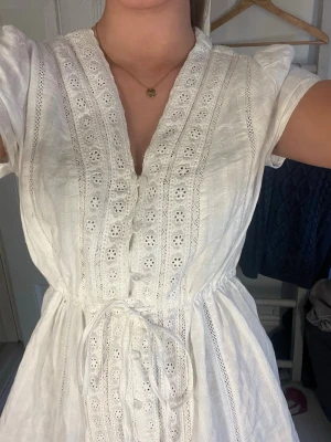 Vit kort klänning med spetsdetaljer - Söt vit kortklänning med v-ringning, knappar framtill och fina spetsdetaljer längs hela klänningen. Klänningen har korta ärmar och ett knytband i midjan för en snygg passform. Perfekt för sommarens alla tillfällen!