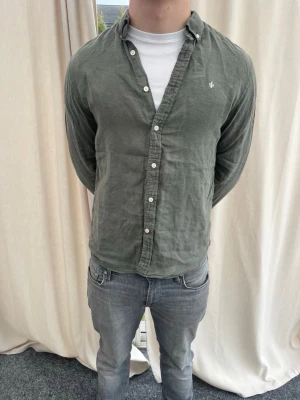 Morris linneskjorta - Stilren grön skjorta med klassisk krage och vita knappar framtill. Skjortan har lång ärm och en liten broderad logga på bröstet. Perfekt för en avslappnad men snygg look.