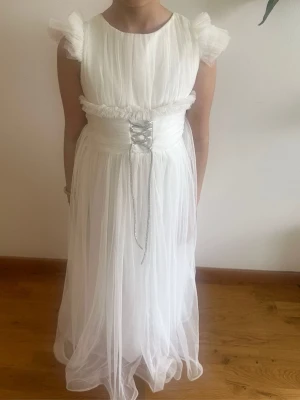 Vit långklänning med snörning - Elegant vit långklänning med transparent tyll och snörning i midjan. Klänningen har korta puffärmar i tyll och ett omlottliknande band runt midjan. Perfekt för speciella tillfällen. Materialet känns lätt och luftigt. Passar 9/10 år.