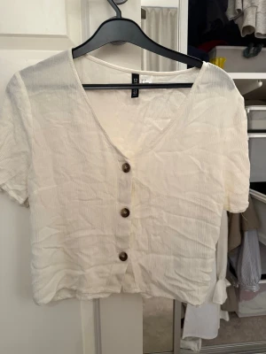 Vit v-ringad blus från H&M Divided - Säljer en vit blus från H&M Divided med v-ringning och tre bruna knappar framtill. Blusen är kortärmad och har en lätt skrynklig struktur som ger en avslappnad vibe. Perfekt att matcha med jeans eller kjol.