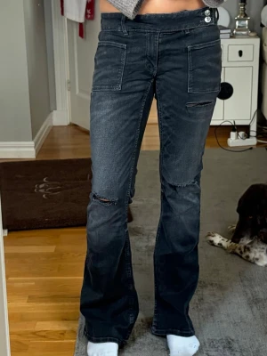 Svarta bootcut jeans med slitningar - Säljer ett par svarta bootcut jeans med coola slitningar på knäna och fickor framtill. Jeansen har en låg midja och knappar i sidan. Perfekta för dig som gillar en avslappnad och trendig look. Märket är gina tricot och dom är i storlek 170 men passar s. Dom är i fint skick, endast använda nån gång.