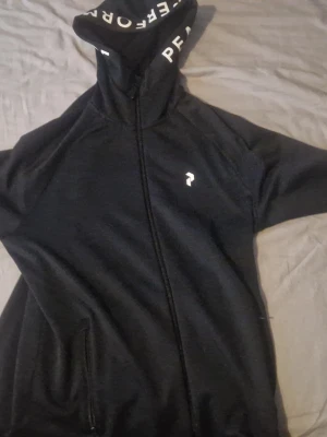 Svart hoodie från Peak Performance - Säljer en svart hoodie från Peak Performance med dragkedja framtill och vit logga på bröstet. Huvan har textdetaljer på insidan och tröjan har fickor på sidorna. Perfekt för en sportig och avslappnad stil.