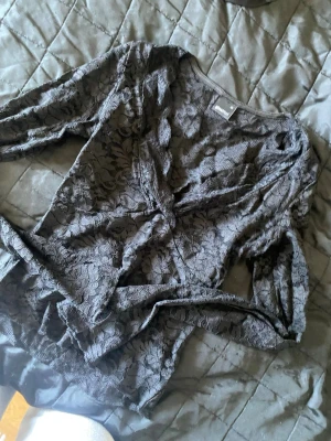 Svart omlottopp i spets - Säljer en svart omlottopp med långärmad design och v-ringning. Toppen är gjord i spetsmaterial med blommigt mönster och har ett knytband i midjan. Perfekt för dig som gillar detaljer och en elegant look.