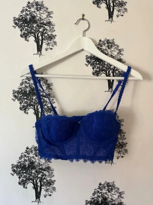 Blå spets-bh med lång modell - Snygg blå bh i lång modell med tunna axelband och vacker spetsdetalj. Bh:n har bygel, vadderade kupor och hakknäppning bak. Spetsen har blommönster och kanten är vågig för en extra fin look.