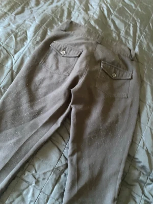 Kostymbyxor med fickor - Snygga kostymbyxor. Byxorna har två bakfickor med lock och knapp, samt bälteshällor och dubbla knappar fram. Perfekta för dig som vill sticka ut lite extra med en diskret men cool detalj.