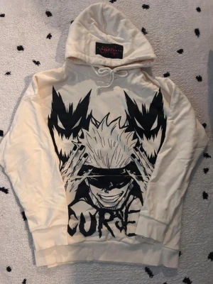 Jujutsu Kaisen Hoodie - Snygg beige hoodie från SKOOT med stort svart anime-tryck framtill som visar Gojo från Jujutsu Kaisen och texten 'CURSE'. Hoodien har huva med dragsko och ribbade muddar vid ärmar och nederkant. Perfekt för dig som gillar streetwear och anime. Custom made, one of a kind hoodie from Korea-based designer SKOOT.