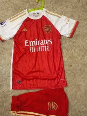 Arsenal matchställ med G.Jesus 9 - Säljer ett Arsenal matchställ från Adidas med G.Jesus och nummer 9 på ryggen. Tröjan är röd med vita ärmar och guldiga detaljer, och har klubbens logga samt sponsortryck. Shorts i rött med guldiga detaljer och Arsenal-emblem. Perfekt för fotbollsfans!