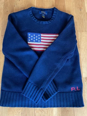 Mörkblå stickad tröja Polo Ralph Lauren - Mörkblå stickad tröja från Polo Ralph Lauren med amerikanska flaggan broderad på bröstet och RL-broderi i rosa vid nederkanten. Rund halsringning och ribbade muddar. Perfekt för dig som gillar klassisk och cool stil.Bara använd ett fåtal gånger och givetvis äkta.