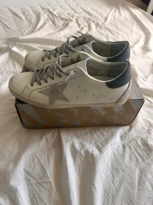 Golden Goose vita sneakers med stjärna - Golden Goose sneakers i vitt läder med grå mockadetaljer och ikonisk stjärna på sidan. Grå skosnören, beige sula och mörkblå hälflik. Klassisk low-top modell med cool vintagekänsla och unika detaljer. Perfekta för dig som gillar streetstyle och trendiga skor. Storlek 45