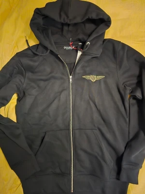 Svart zip-hoodie från Gul&Blå - Cool svart hoodie med dragkedja från Gul&Blå. Den har huva, fickor fram och snyggt broderat G&B-emblem i gult både på bröstet och ryggen. Perfekt för dig som gillar streetstyle och vill ha något bekvämt och stilrent. Materialet är mjukt och känns skönt mot huden.