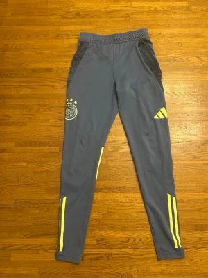 Ajax träningsbyxor från Adidas - Snygga blå träningsbyxor från Adidas med Ajax Amsterdam-logga och gula detaljer. Byxorna har smal passform, elastisk midja och sidofickor med svarta paneler. Perfekta för fotbollsträning eller gymmet.