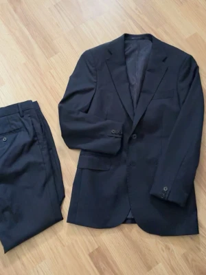 Mörkblå kostym från These Glory Days - Klassisk mörkblå kostym från These Glory Days med enkelknäppt kavaj och matchande byxor. Kavajen har smal passform, två knappar och fickor med lock. Perfekt för dig som vill ha en stilren och tidlös look. Knappt använd. Omsydd vid ärmarna. Nypris ligger på 2400 så klaga gärna inte på priset