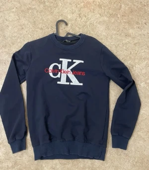 Mörkblå sweatshirt Calvin Klein - Mörkblå sweatshirt från Calvin Klein Jeans med stor vit CK-logga och röd text på bröstet. Tröjan har rund halsringning, ribbade muddar vid ärmslut och nederkant samt en klassisk passform. Perfekt för en avslappnad och trendig look.