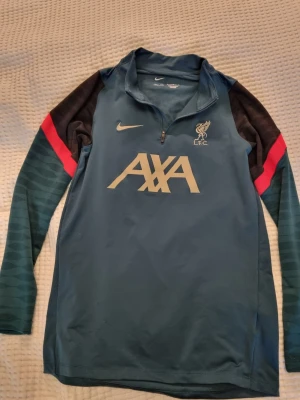 Nike Liverpool långärmad träningströja - Långärmad träningströja från Nike för Liverpool FC i grönt med svarta och röda detaljer på ärmarna. Tröjan har halv dragkedja, Dri-FIT-material och stora tryck med AXA och Liverpool-logga på bröstet. Fast pris, inga byten.