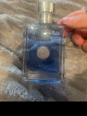 Versace Pour Homme Eau de Toilette 100 ml - Versace Pour Homme Eau de Toilette med elegant, rektangulär glasflaska i blått och silverfärgat lock. Flaskan har Versaces ikoniska Medusa-symbol i mitten. Medföljer original svart kartong med silverdetaljer och klassisk design.