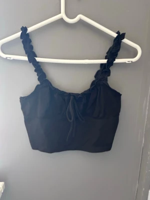 Svart croppad linnetopp med volangband - Snygg svart croppad linnetopp med volangdetaljer på axelbanden och knyt framtill. Enkel att matcha.