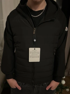 Svart Moncler pufferjacka med huva - Säljer en svart pufferjacka från Moncler med huva och två dragkedjeförsedda fickor. Jackan har Moncler-logga på ärmen och quiltad framsida. Stängs med dragkedja och har en clean, modern look som passar perfekt nu när våren är här. Jackan är i storlek M men kan nog även passa i S. För fler bilder ba kontakta DM.