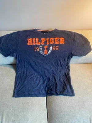 Marinblå Hilfiger t-shirt med tryck - Snygg marinblå t-shirt från Tommy Hilfiger med orange text och emblem på bröstet. Trycket visar 'HILFIGER', '1985' och ett sköldmärke. Klassisk passform och rund hals. Perfekt till jeans eller shorts.