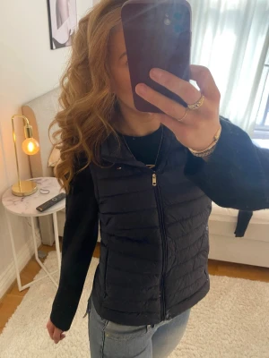 Mörkblå väst från Tommy Hilfiger - Snygg mörkblå dunväst från Tommy Hilfiger med dragkedja framtill och huva. Västen har en quiltad design och en liten logga på bröstet. Perfekt att slänga över en hoodie eller tröja för en casual look.