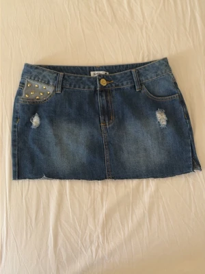 Blå jeanskjol med nitar och slitningar - Säljer en blå jeanskjol i minilängd med slitna detaljer och rå kant. Kjolen har nitar på ena fickan framtill och klassiska fickor både fram och bak. Perfekt för dig som gillar en edgy och trendig stil.