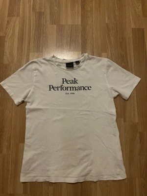 Vit Peak Performance t-shirt - Klassisk vit t-shirt från Peak Performance med svart logotyp och text 'Est. 1986' på bröstet. T-shirten har rund hals och korta ärmar, perfekt för en clean och enkel stil. Tillverkad i mjuk bomull för skön komfort. Den har ett litet hål på axeln men inget man märker mycket
