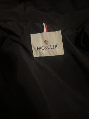 MONCLER WINDBREAKER  - Snygg svart dunjacka från Moncler med klassisk logotyp på insidan och detaljer i rött, vitt och blått. Jackan är tillverkad i slitstarkt material och fylld med dun för maximal värme under kalla vinterdagar. Perfekt för dig som vill ha både stil och funktion.