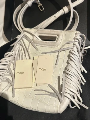 Vit handväska med fransar från Maje - Säljer en vit handväska från Maje i croco-mönstrat konstläder. Väskan har dekorativa fransar på sidorna, ett handtag upptill och både axelrem i läder och kedja. Perfekt accessoar för att lyfta din outfit med en trendig touch.