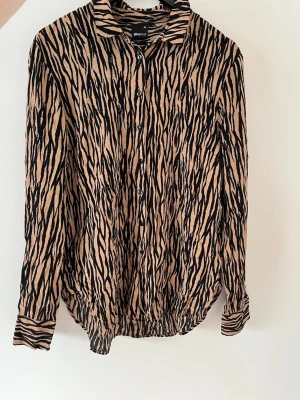 Skjorta - Snygg mönstrad skjorta från Gina Tricot i brunt/beige och svart. Skjortan har klassisk krage, knappar framtill och långa ärmar. Tillverkad i ett lätt och mjukt material som känns skönt mot huden. Fint skick! 