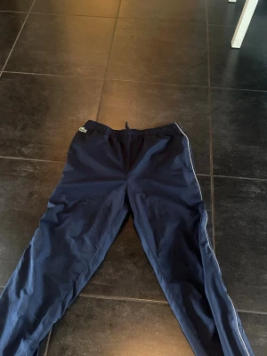 Mörkblå mjukisbyxor med logga - Säljer ett par mörkblå mjukisbyxor från Lacoste med vit piping längs sidorna och elastisk midja. Perfekta för chill eller träning, med diskret logga på höften. Materialet är lätt och bekvämt.