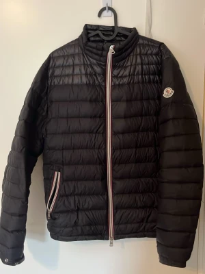 Svart dunjacka från Moncler - Snygg svart dunjacka från Moncler med quiltad design och raka ärmar. Jackan har dragkedja framtill med Monclers signaturfärger och en matchande dragkedja på fickan. Märkeslogga på vänster ärm. Perfekt för kalla vinterdagar.