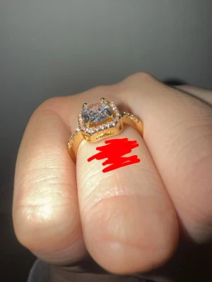 Guldfärgad ring med stor sten - Snygg guldfärgad ring med en stor, fyrkantig klar sten i mitten och små gnistrande stenar runt om. Ringen har en elegant och lyxig känsla med detaljerade infattningar. Perfekt för dig som vill sticka ut med accessoarerna.