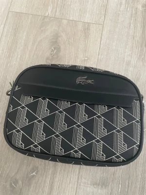 Svart axelväska från Lacoste - Snygg svart axelväska från Lacoste med grafiskt mönster i vitt och silverfärgad krokodillogga framtill. Väskan har en rektangulär form och är tillverkad i slitstarkt syntetmaterial. Perfekt för att bära dina viktigaste grejer med stil.