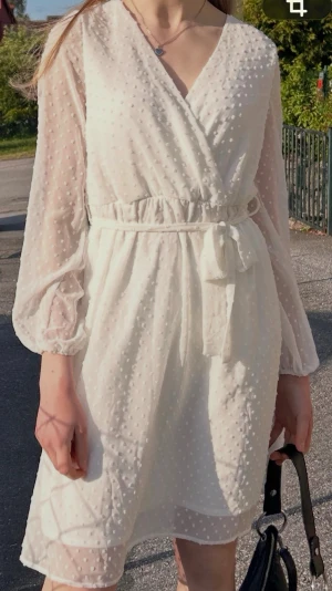 Vit klänning, avslutning, student  - Vit V-ringad, långärmad klänning. Vita prickar med knytning i midjan, dragkedja i ryggen. Når till knäna på mig som är 173cm lång. Helt oanvänd med lappen kvar, nypris: 499:-.  Material: 100% Polyester. 