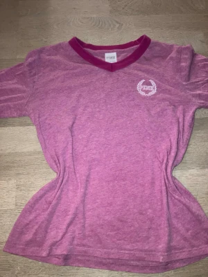 Rosa V-ringad T-shirt från PINK - Snygg rosa T-shirt från PINK med v-ringad hals och kontrasterande mörkrosa kant. Liten vit logga med lagerkrans på bröstet. Perfekt för dig som gillar en enkel men ändå trendig look. Skön och mjuk i materialet.