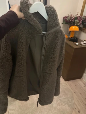 Mörkbrun teddyjacka med dragkedja - Mysig mörkbrun teddyjacka med hög krage och dragkedja framtill. Jackan har två sidofickor och är gjord i fluffigt, mjukt material som håller dig varm. Perfekt för kyliga dagar och ger en avslappnad, trendig look.