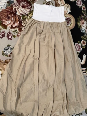 Beige långklänning med vit överdel - Säljer en beige långklänning med vit, axelbandslös överdel. Kjoldelen är vid och har ett skirt, luftigt tyg med veck. Perfekt för sommarens alla tillfällen. Märke: ZARA.
