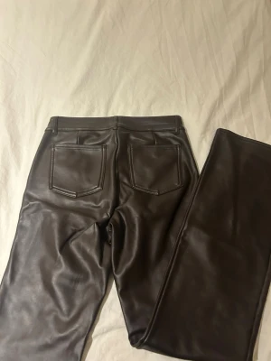 Bruna fuskskinnjeans med raka ben - Säljer ett par bruna jeans i fuskskinn med raka ben och klassisk femficksmodell. Jeansen har bälteshällor, knappstängning och en snygg glansig finish som ger en cool look. Perfekta för dig som vill sticka ut lite extra.